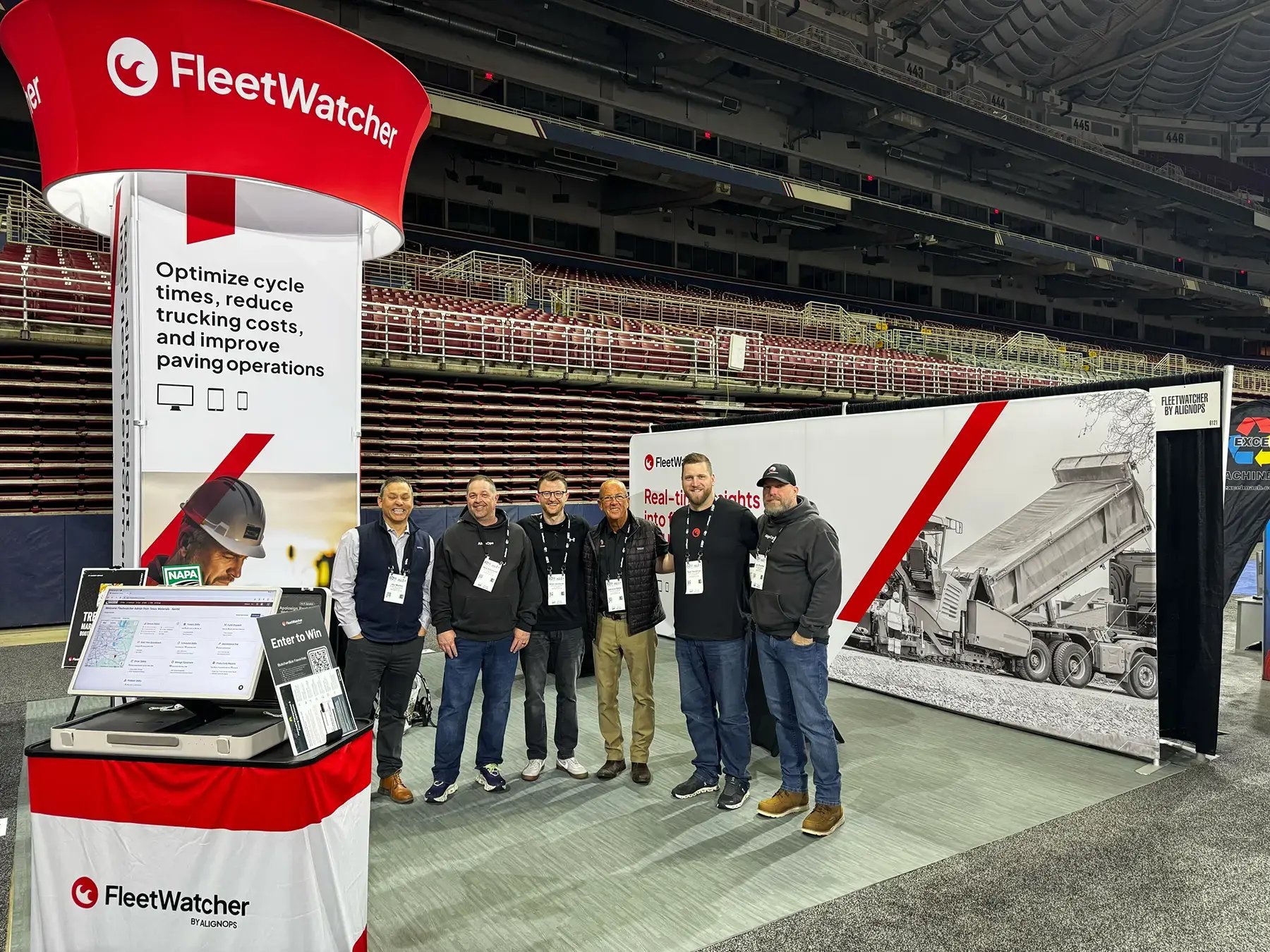 A group of six people stands in front of a FleetWatcher booth at a trade show