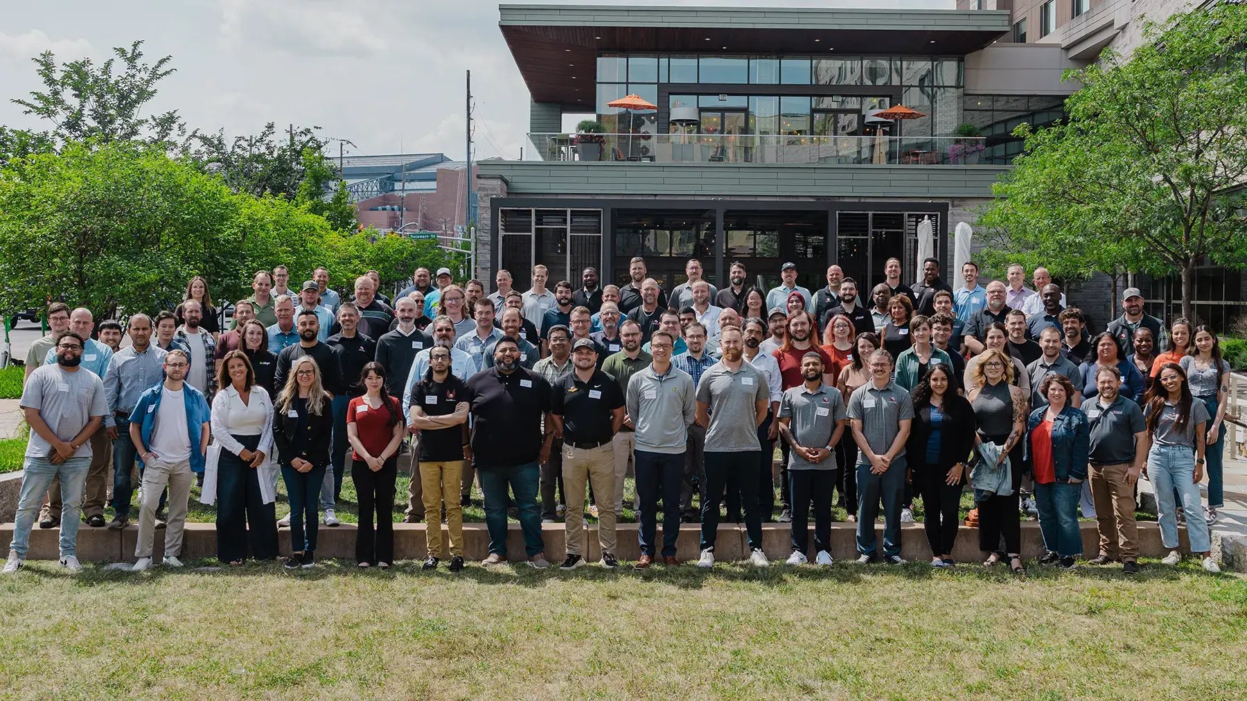 A large group of diverse people stands smiling outdoors, with a modern building and greenery in the background, conveying a cheerful and united atmosphere