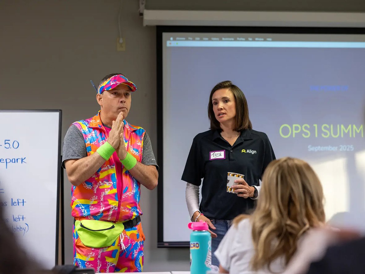 A man in vibrant, colorful athletic wear stands with hands clasped beside a woman in a casual black shirt, addressing an audience at a presentation
