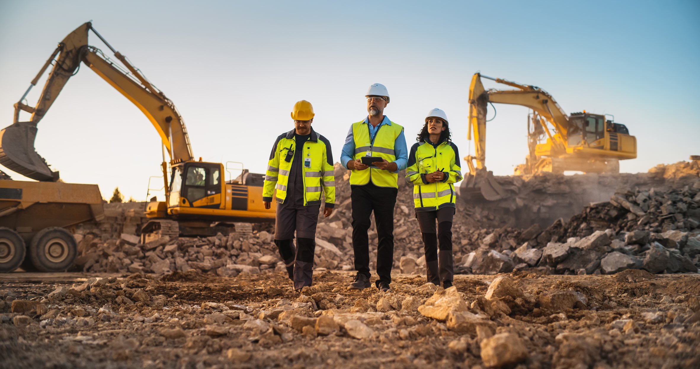 construction professionals walking throuhg a dirt job site with large construction diggers in the background