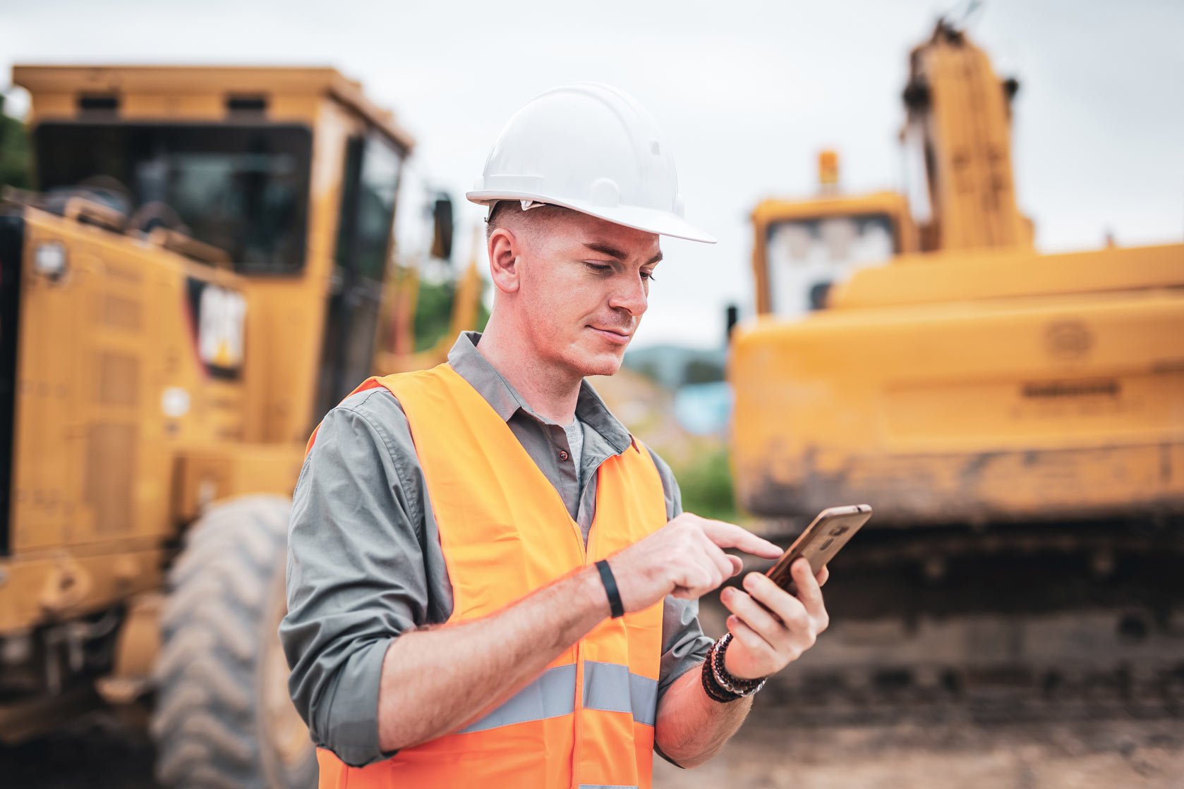 project manager standing near large construction machinery while tapping mobile phone